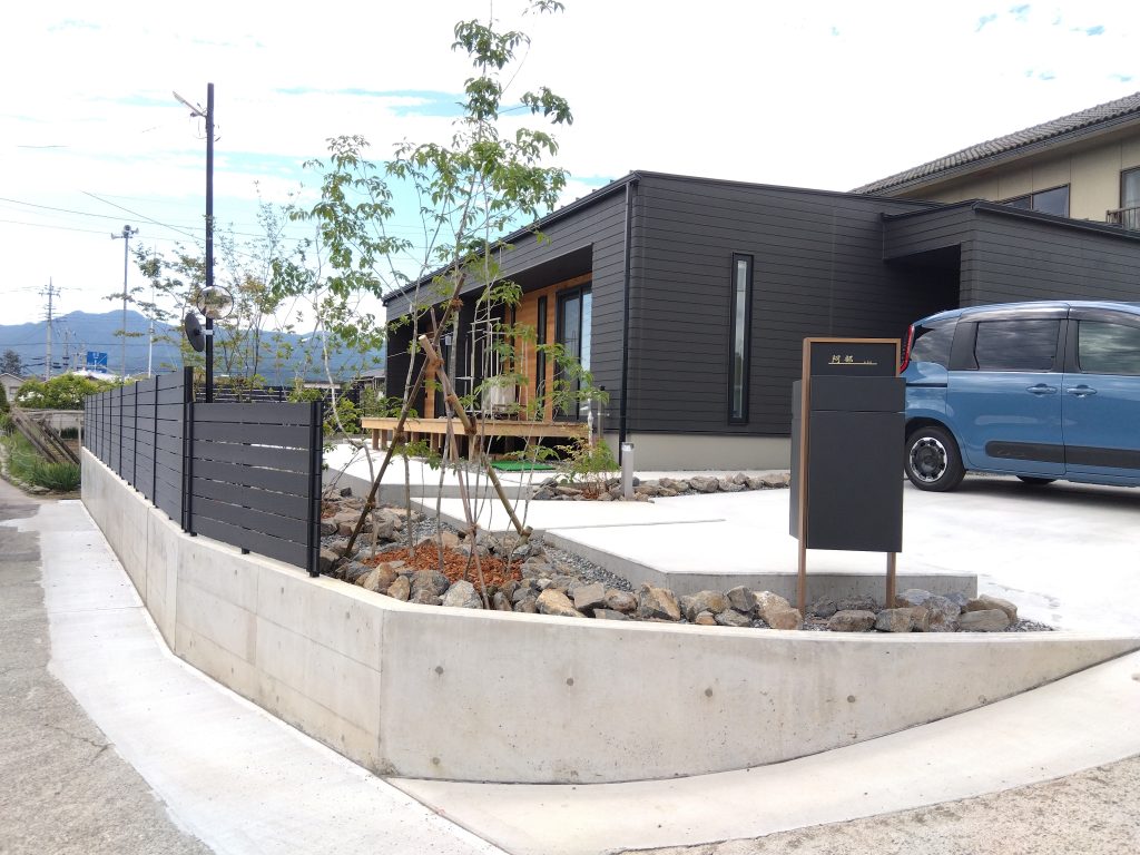 山梨県山梨市の外構施工例。黒い建物に合わせた黒色人工木材フェンスとLIXIL機能門柱、人工芝と雑木が映えるモダンエントランス。