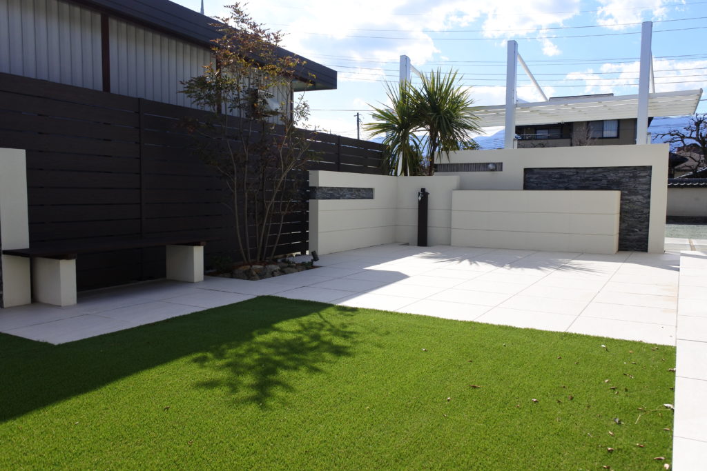 山梨県甲府市の外構施工例。高級感のある大判タイル門柱と、その脇を彩る雑木の植栽、割栗石が調和したナチュラルモダンなアプローチ。