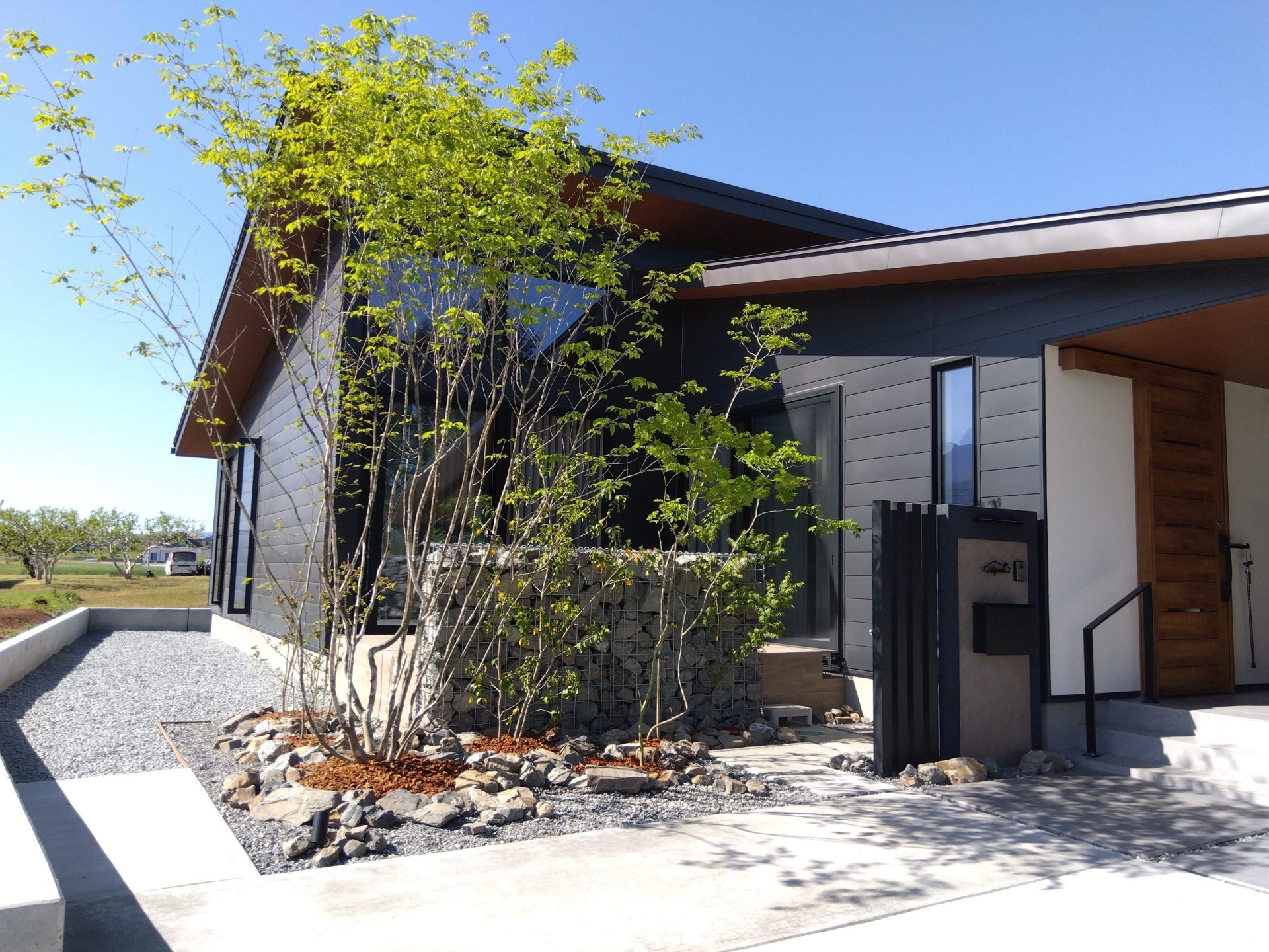 山梨県南アルプス市の外構施工例。蛇篭（ガビオン）と黒タイル貼りの意匠壁、雑木の庭が調和したナチュラルモダンなエントランス。
