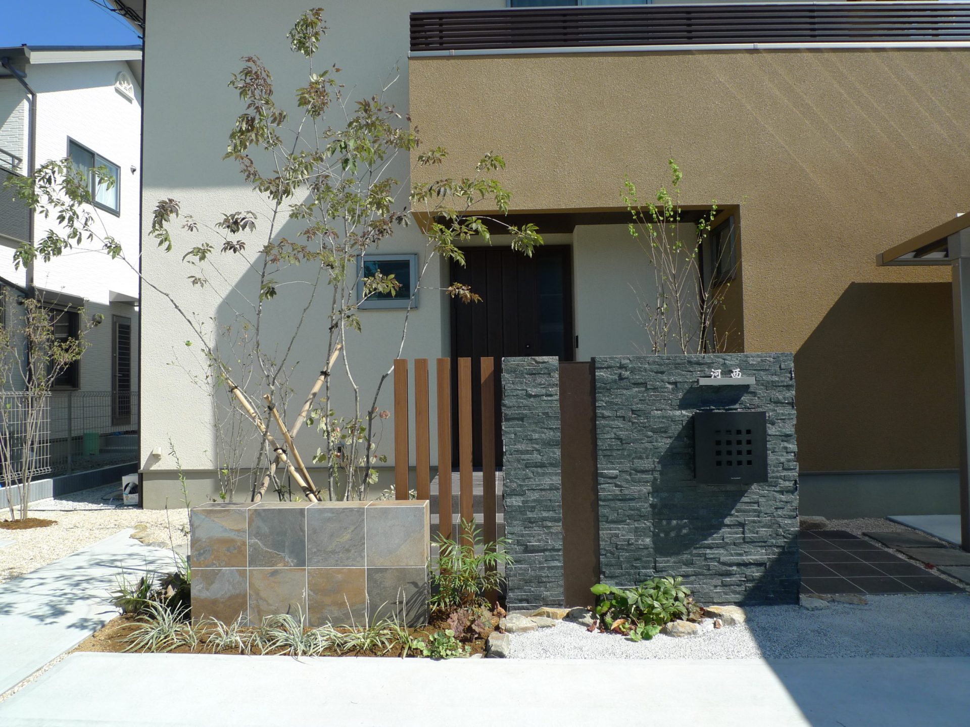 山梨県甲府市の外構施工例。グレーの大判タイル門柱と、スタイリッシュなカーポート、周囲を彩る雑木の植栽が調和したナチュラルモダンな外構。
